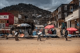 Busy street market with colorful umbrellas and people walking, set against a backdrop of hills and overcast skies.
