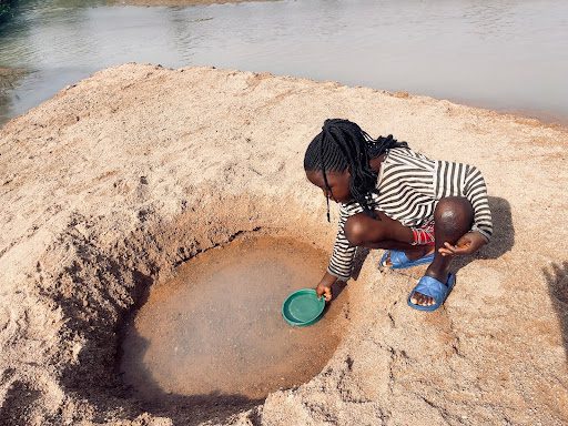 Child collecting water from a shallow hole near a riverbank, wearing a striped shirt and sandals, using a green container.