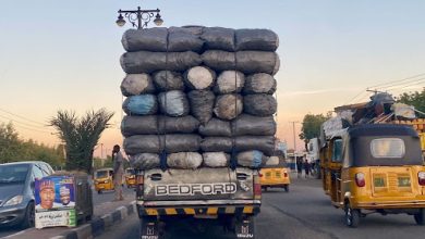 Truck overloaded with bags drives on a busy street at sunset, accompanied by auto rickshaws and passing vehicles.