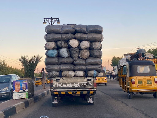 Truck overloaded with bags drives on a busy street at sunset, accompanied by auto rickshaws and passing vehicles.