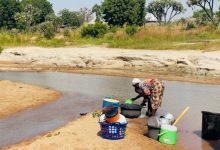 Person washing dishes in a shallow river, surrounded by buckets and basins, with trees and greenery in the background.