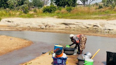Person washing dishes in a shallow river, surrounded by buckets and basins, with trees and greenery in the background.