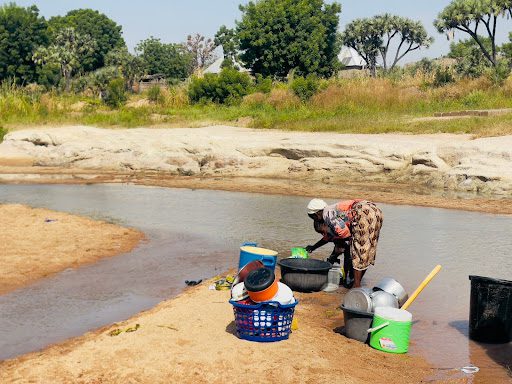 Person washing dishes in a shallow river, surrounded by buckets and basins, with trees and greenery in the background.