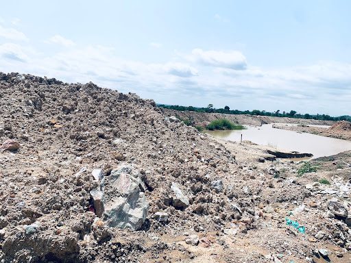 Rocky terrain with piles of dirt beside a small body of water under a cloudy sky.