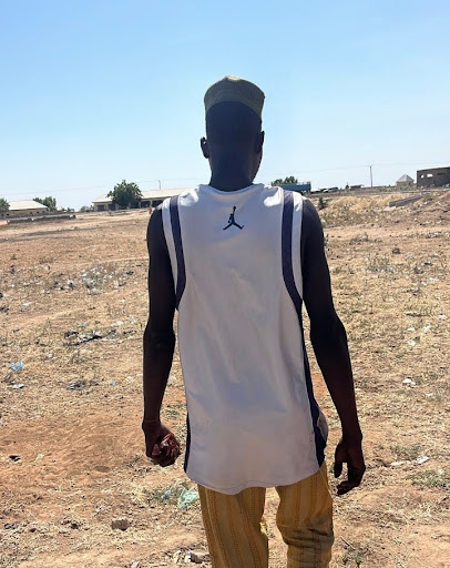 Man in a white sleeveless shirt and striped pants walks on a dry field under a clear blue sky, with buildings in the background.