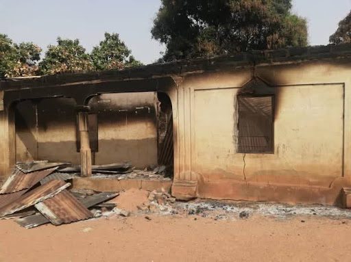 A burned, partially collapsed building with debris on the ground, surrounded by trees.