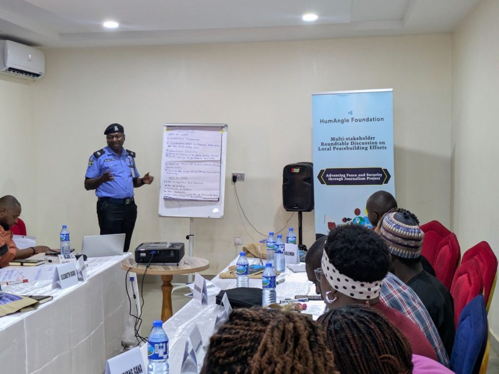 A police officer presents at a workshop, surrounded by participants. A banner reads "HumAngle Foundation" and relates to peacebuilding efforts.