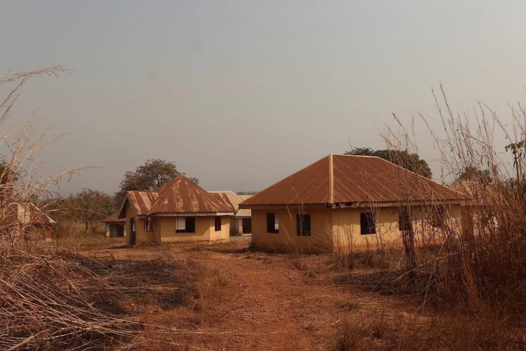 Two small, yellow houses with rusty red roofs in a dry, grassy, rural landscape under a clear sky.