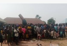 A large group of people, including children, standing outdoors near buildings in a rural setting.