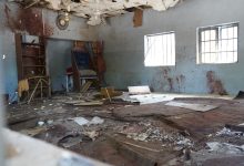Dilapidated room with debris, broken ceiling, and stained walls. Two barred windows let in light.