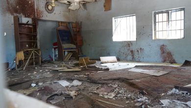 Dilapidated room with debris, broken ceiling, and stained walls. Two barred windows let in light.