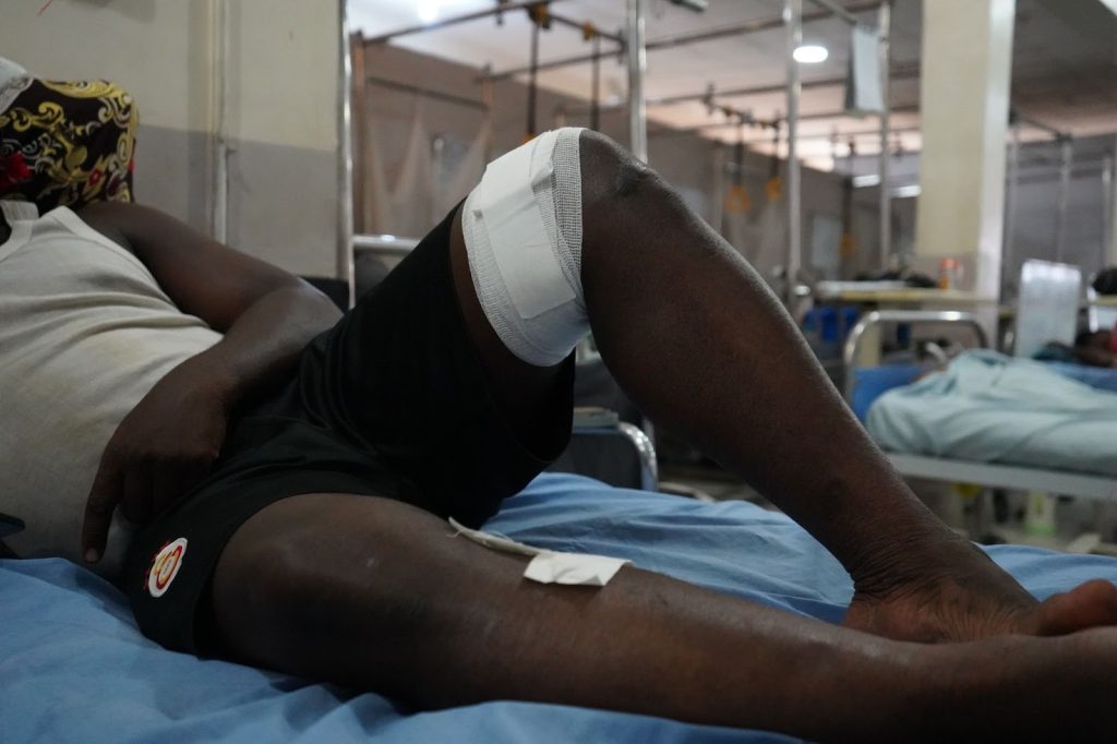 A person with a bandaged knee lies on a hospital bed, resting in a ward with other occupied beds in the background.