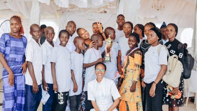 A group of people smiling, holding a trophy, indoors. Some wear matching gray shirts, others colorful attire, with a white draped backdrop.