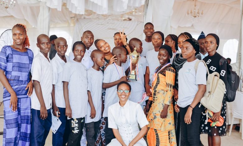 A group of people smiling, holding a trophy, indoors. Some wear matching gray shirts, others colorful attire, with a white draped backdrop.