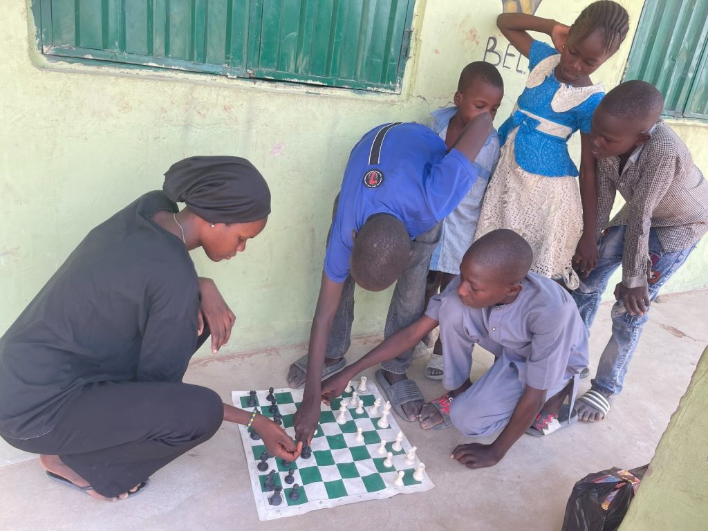 A group of children and an adult play chess on a green and white board, huddled closely in front of a green wall.