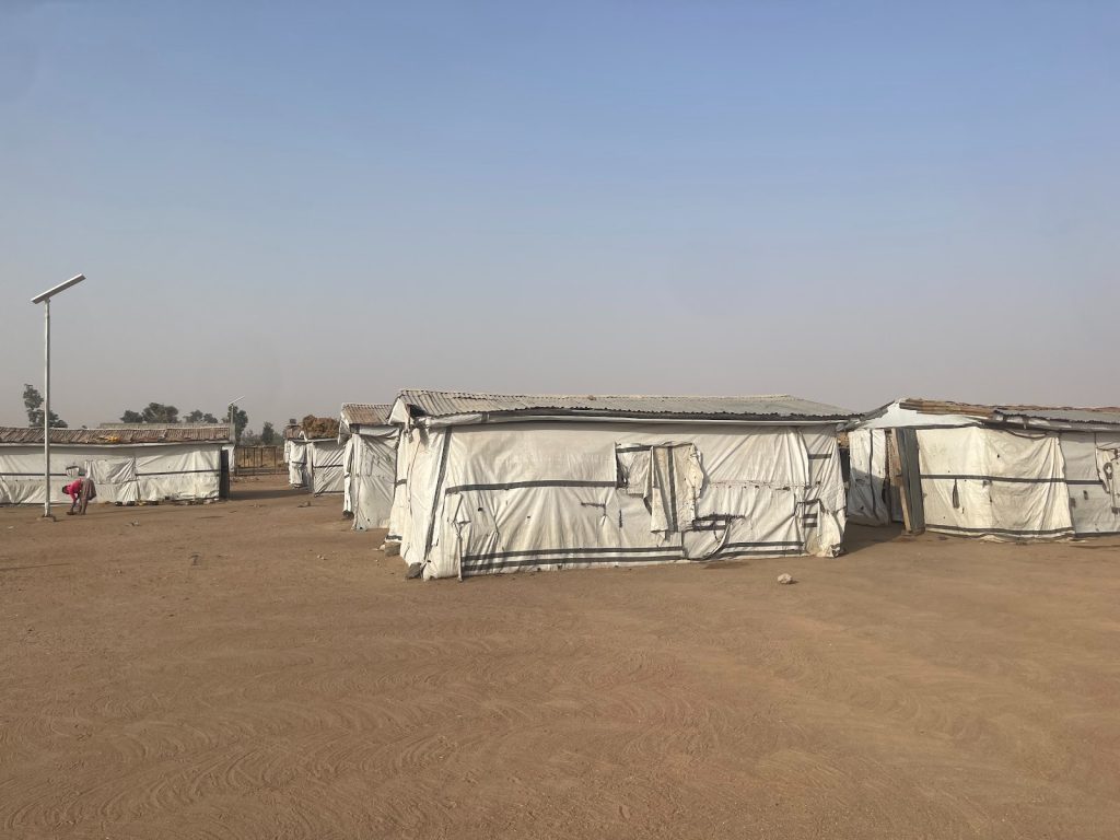 Rows of white tents under a clear blue sky in a dry, dusty area, with a solar light pole and a child in the background.