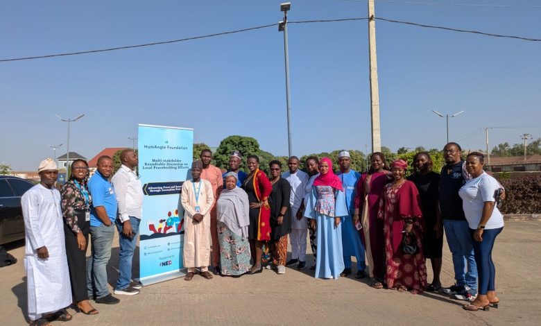 A group of people standing together outdoors near a banner for a multi-stakeholder roundtable on local peacebuilding efforts.