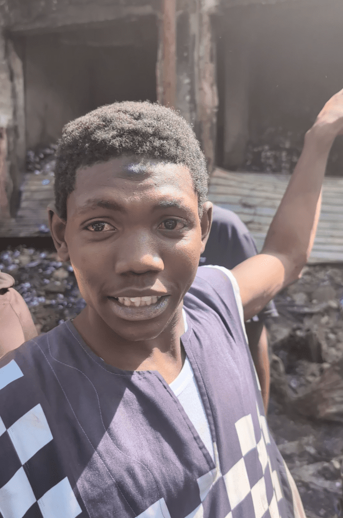 Smiling person taking a selfie outdoors, arm raised, with a blurred background of a building.