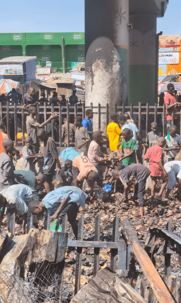 People sorting through debris near a fence, surrounded by makeshift structures under a bridge.