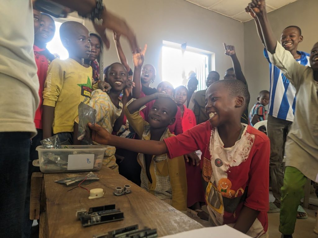 Children excitedly gather around a table with electronics parts, eagerly learning and engaging in a hands-on activity.