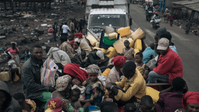 A crowded truck carries people with belongings, including jerry cans, down a road, while others walk nearby in a busy, makeshift setting.