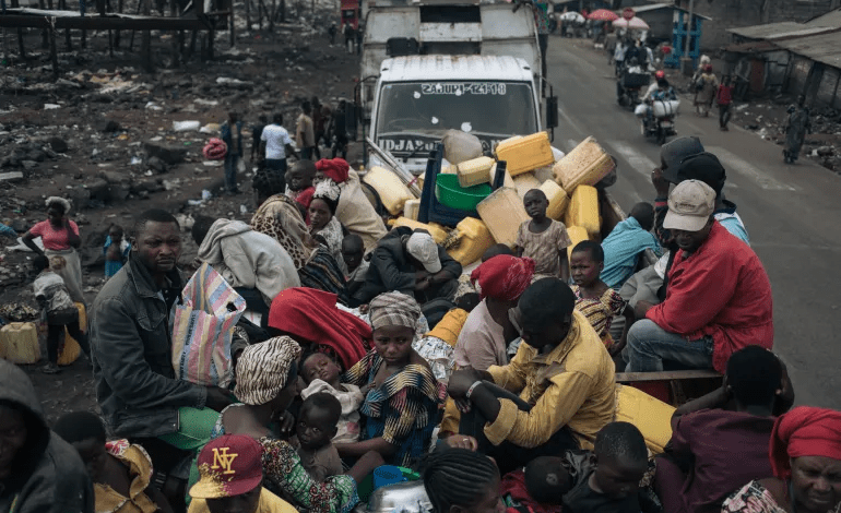 A crowded truck carries people with belongings, including jerry cans, down a road, while others walk nearby in a busy, makeshift setting.