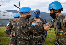 UN peacekeepers in blue helmets share an elbow bump beside a UN helicopter, set against a backdrop of green hills.