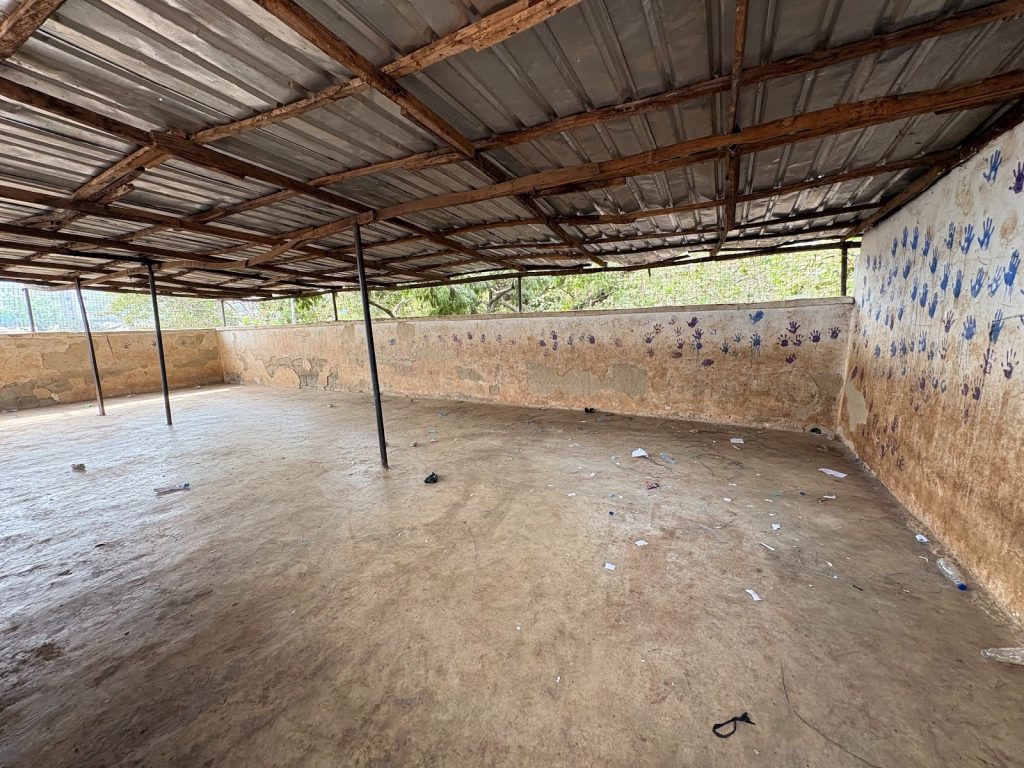 Covered concrete space with metal roof, support beams, and painted handprints on walls. Scattered debris on floor, open view to greenery.