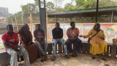 A group of people sitting on benches outdoors, with colorful walls and trees in the background.