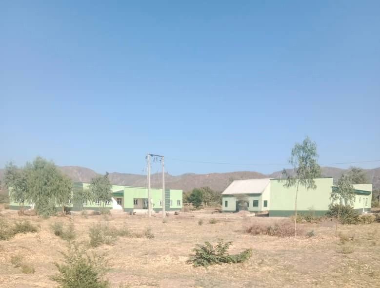 Green buildings under a clear blue sky, with dry grass and scattered trees in the foreground. Hills are visible in the background.