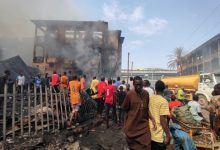 Crowd gathers around a smoke-filled, fire-damaged building as a tanker arrives; debris lies scattered on the ground.