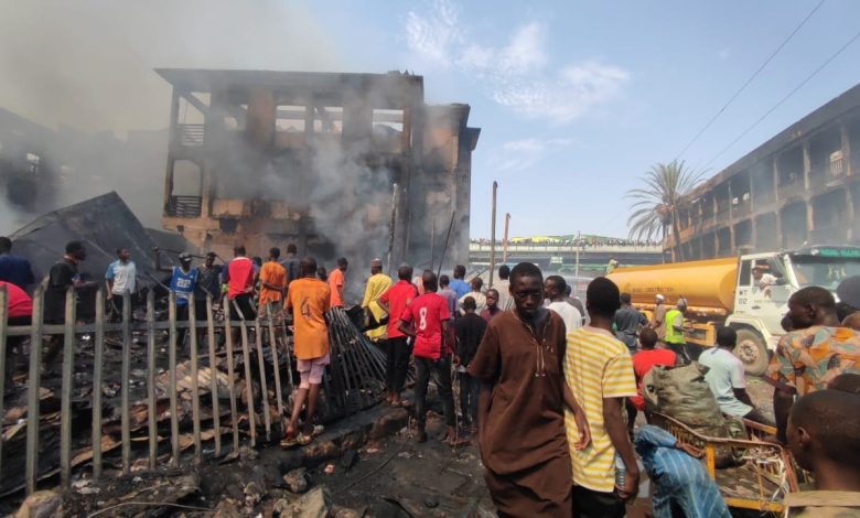 Crowd gathers around a smoke-filled, fire-damaged building as a tanker arrives; debris lies scattered on the ground.