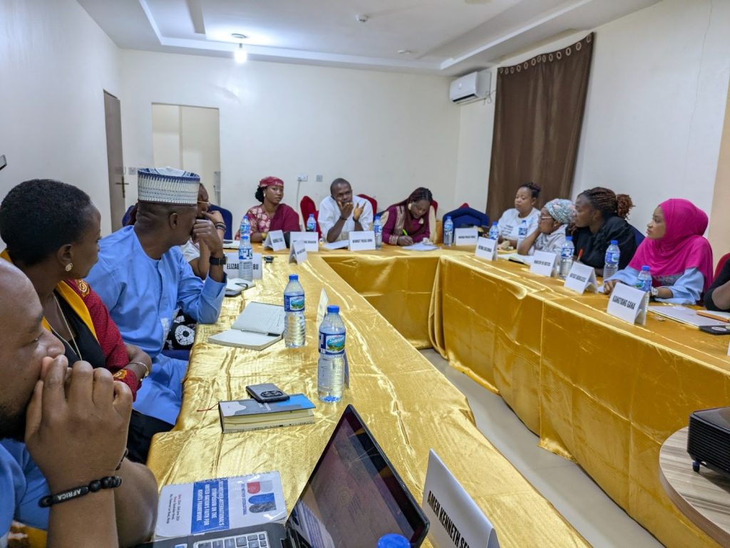 People sitting around a table in a meeting room, engaged in discussion. Nameplates and notepads are visible on the table.