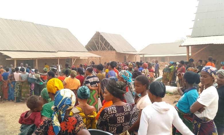 A large group of people gathered outside brick buildings with corrugated metal roofs, many holding containers and wearing colorful clothing.