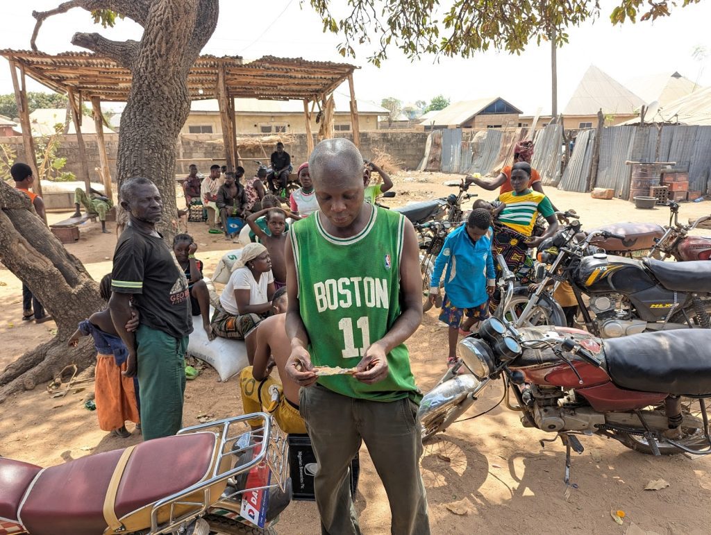 People gather under a tree near parked motorcycles in a rural setting, engaging in various activities.