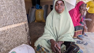 A woman in a green dress and headscarf sits outside a building, with colorful sacks of grain behind and two women sitting nearby.
