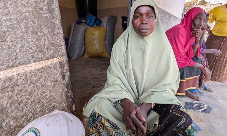 A woman in a green dress and headscarf sits outside a building, with colorful sacks of grain behind and two women sitting nearby.
