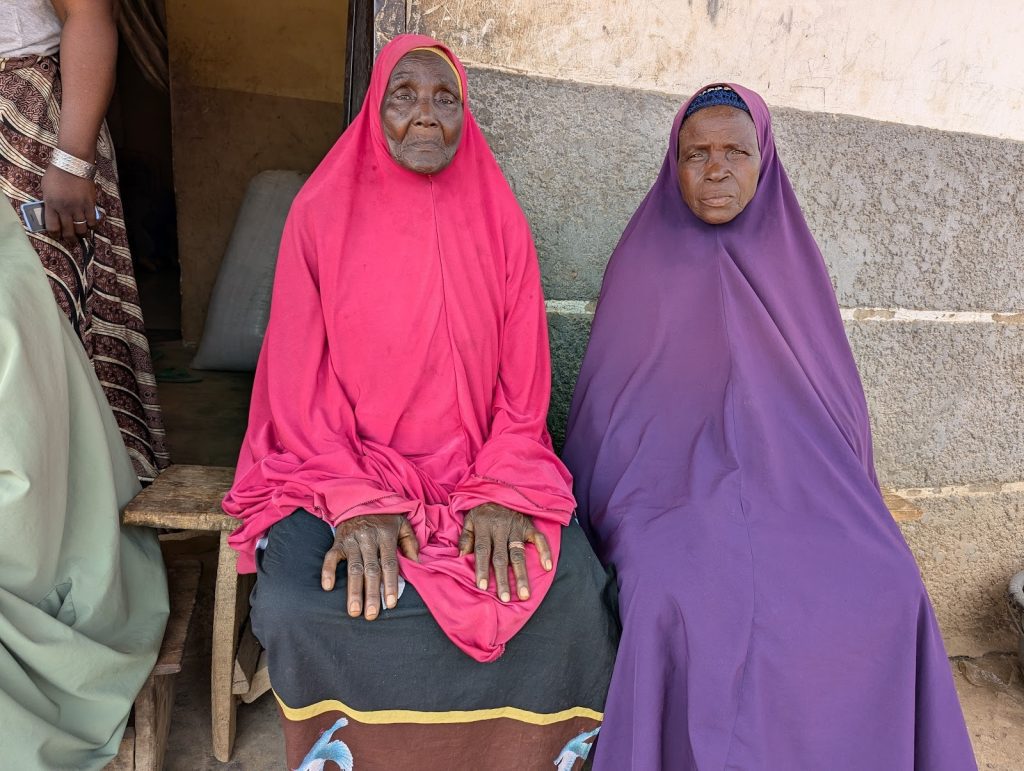 Two women in vibrant pink and purple hijabs sit side by side on a bench against a textured concrete wall.