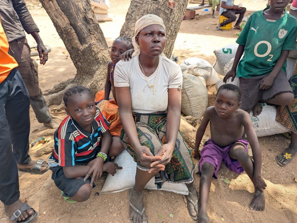 A woman and three children sit on sacks under a tree, surrounded by people in a sunny outdoor setting.