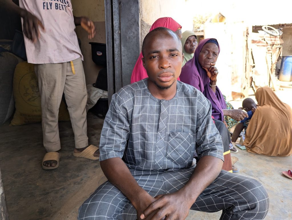 Man in patterned outfit sitting in front of a doorway, with people in colorful attire sitting and standing nearby.