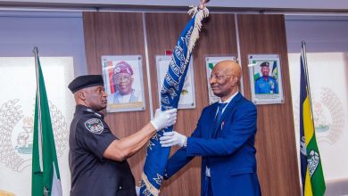 Two men exchange a decorated staff in an official setting, with flags and portraits in the background.
