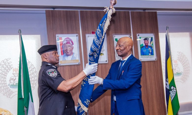 Two men exchange a decorated staff in an official setting, with flags and portraits in the background.
