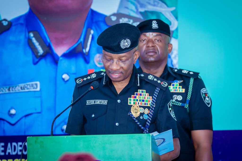 Two uniformed police officers stand at a podium. The foreground officer reads from a paper, wearing a decorated uniform and cap.