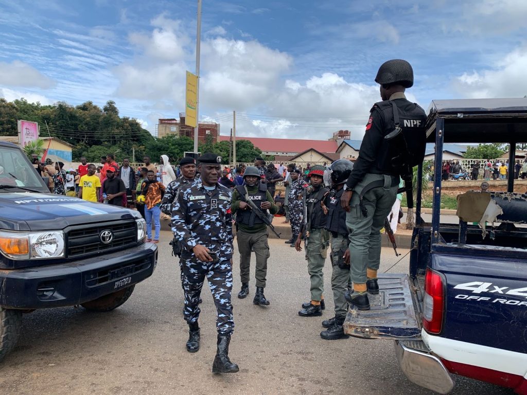 Police officers stand near vehicles and a crowd on a street, with trees and buildings in the background under a cloudy sky.