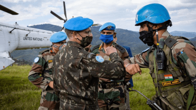UN peacekeepers in blue helmets share an elbow bump beside a UN helicopter, set against a backdrop of green hills.