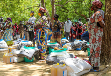 People in colorful clothing stand by supplies, including jugs and bags, under trees in an outdoor setting.