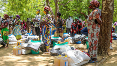 People in colorful clothing stand by supplies, including jugs and bags, under trees in an outdoor setting.