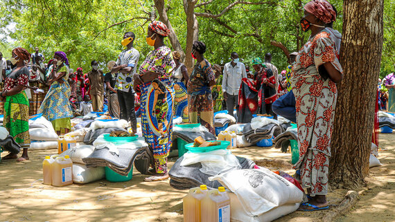 People in colorful clothing stand by supplies, including jugs and bags, under trees in an outdoor setting.