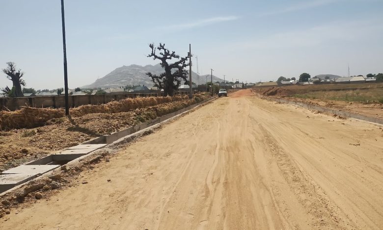 A dusty rural road with sparse trees, a parked car, and distant hills under a clear blue sky.