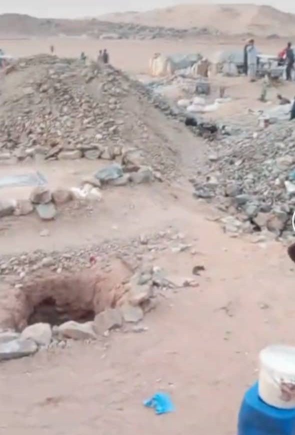 Desert landscape with stone mounds, scattered rocks, and distant people near tents.